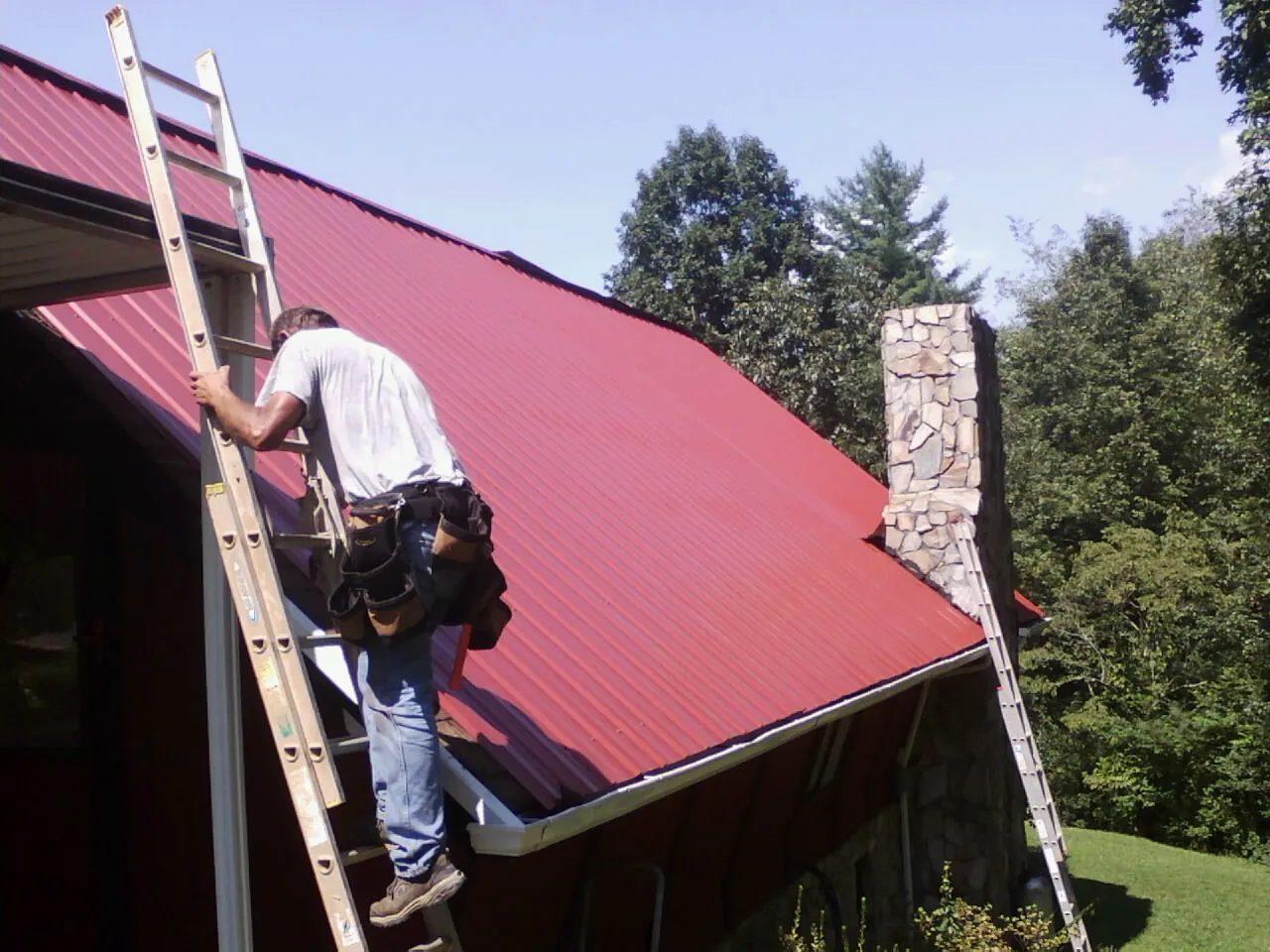 Roofer on a ladder beside a red metal roof and stone chimney — Soffit & Fascia Repair by Roofing Brothers in Belt